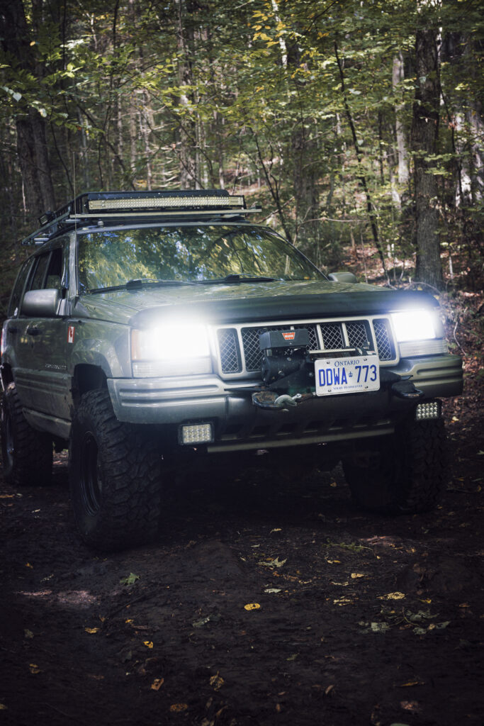 A sliver grey Jeep Cherokee head on in a forest. Lights are bloomed out.