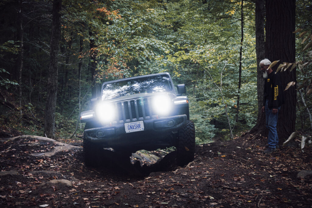 A grey Jeep Gladiator is climbing a hill, tires straddling a v-notch rock formation in a forest.